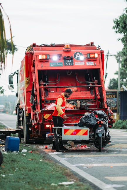 A worker wearing a high-visibility yellow and orange vest is operating a large red waste collection vehicle on a street. The back of the vehicle is open, revealing mechanical components and a compactor. The worker is standing slightly to the side, handling a black rubbish bag placed on a small metal platform attached to the vehicle. Nearby, there is a motorcycle parked close to the curb, with its seat and side panels visible. The surrounding environment includes a grassy verge, a sidewalk, and a few trees in the background, with utility poles and power lines overhead. The sky appears overcast, casting diffused light over the scene. This image depicts an on-site waste collection operation, which is typically part of private rubbish removal services such as those offered by Waste Removal Mayfair, emphasizing efficient rubbish clearance in an urban setting.