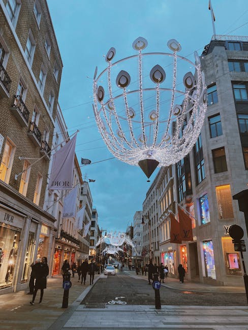 A wide city pedestrian street during twilight, with numerous shoppers walking along the paved walkway lined with luxury retail stores displaying illuminated window displays. Large decorative streetlights shaped like oversized spherical chandeliers, with black and white teardrop-shaped accents, hang overhead, casting a soft glow on the scene. Surrounding buildings are multi-storey commercial structures with modern glass and stone facades; some show signs of retail branding, including a Dior storefront on the left. Pedestrians are seen in coats and jackets, some carrying shopping bags, creating an active urban atmosphere. The sky above is a clear blue, indicating early evening. The scene exhibits a festive or decorative ambiance, with bright, reflective surfaces and clean, well-maintained surroundings, aligning with an area that prioritizes private or independent retail and street-level decorative lighting. This detailed environment underscores a high-end shopping district often associated with independent aesthetic appeal and exclusive retail experiences, consistent with luxury shopping atmospheres rather than typical rubbish removal contexts, but the scene’s cleanliness supports the notion of regular waste management or tidy urban upkeep by local services like Waste Removal Mayfair.