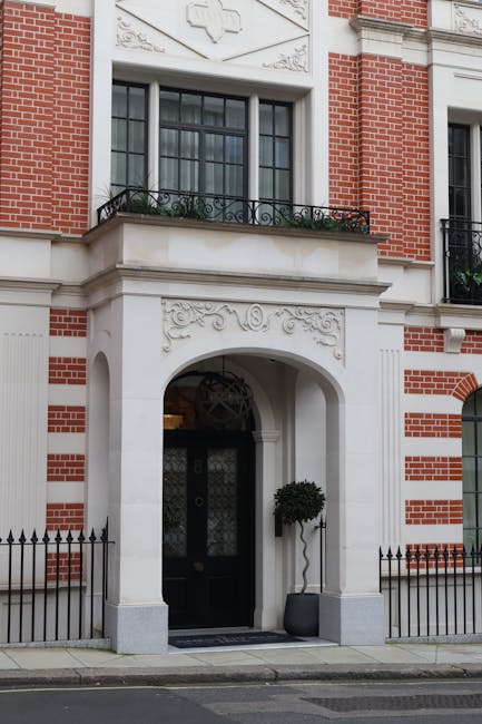 The image shows the entrance of a historic or period-style building with a combination of red brick and light-colored stone or plaster detailing, characteristic of traditional London architecture. The doorway is framed by a white arch with intricate decorative carvings above it, leading to a black door with a glass panel and an ornate metal grille. To the right of the entrance, there is a small potted plant with a neatly trimmed, spherical topiary placed in a dark, rounded container, adding a touch of greenery to the otherwise structured façade. Above the arch, a large rectangular window with multiple smaller panes is visible, framed by the same light-colored material, and features a black wrought-iron balcony with a simple, elegant design, decorated with a few plants or flowers in small containers. The building’s exterior displays a detailed pattern of red brickwork interspersed with white stone accents, and the surrounding pavement is clean, with a short black metal fence partially visible on either side of the doorway, indicating a private property or estate. The natural daylight illuminates the scene softly, emphasizing the architectural details and the clean, well-maintained appearance, reflecting the context of private residential or estate clearance services often handled by waste management specialists such as Waste Removal Mayfair.