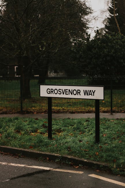 A rectangular white street sign with black uppercase lettering reading 'GROSVERNOR WAY' is mounted on two black metal posts situated on a grassy verge beside a paved road. The grass appears slightly damp with scattered fallen leaves, indicating a recent rain or autumnal season. Behind the sign, there is a black metal fence extending horizontally, which encloses an area with a mix of leafless trees and bushy green foliage, suggesting a park or garden setting. Above, the sky is overcast with grey clouds, providing diffuse, subdued lighting that emphasizes the textures and colors of the natural environment. In the context of private waste management or alternative rubbish collection services, this street scene with the sign and greenery could be part of a residential area where such services are often arranged privately. The scene's simplicity and focus on the street sign and its immediate surroundings highlight the typical outdoor environment associated with urban or suburban rubbish removal and estate clearance operations, as coordinated by companies like Waste Removal Mayfair.