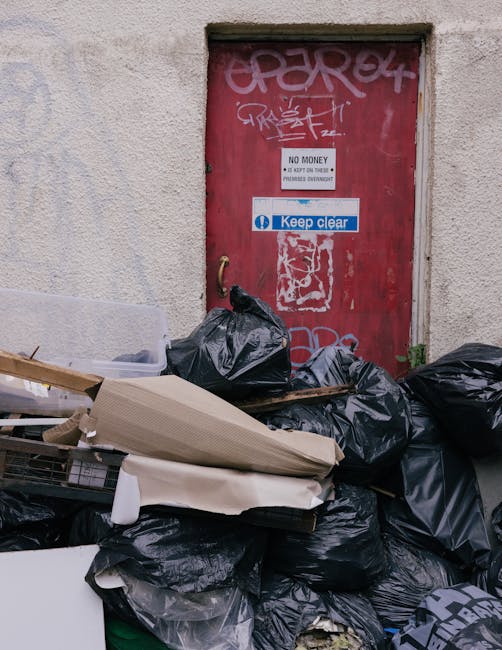 A pile of black garbage bags filled with waste material stacked against a weathered, beige exterior wall of a building. The bags are situated at the base of a red metal door, which is covered in graffiti tags and stickers, including a prominent blue and white 'Keep clear' sign. Some cardboard boxes and discarded packaging materials are partially visible among the rubbish, with a small plastic container on the left side. The door has a brass handle and is set within a frame that appears to be slightly recessed. The surrounding wall shows signs of wear and age, with a rough texture and uneven coloration. The overall scene suggests a private waste collection area, managed by Waste Removal Mayfair, relating to alternative rubbish removal or on-site clearance services typical of Mayfair's urban environment, and highlights the importance of proper waste disposal and junk removal solutions in busy city areas.