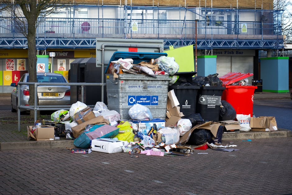 A crowded collection of various rubbish bags, cardboard boxes, and loose waste items piled around a large grey mixed waste bin on a paved urban sidewalk. The bin has a blue label indicating it is for mixed paper and card, with some trash spilling over its open top, including crumpled paper, cardboard, and plastic packaging. Surrounding the bin, additional black and red rubbish bags, flattened boxes, and smaller debris are scattered across the paving stones. Behind the waste area, a grey car with a yellow license plate is parked parallel to a metal railing that surrounds some young trees planted along the pavement. In the background, there are commercial storefronts with brightly colored signage, and a building under construction with blue scaffolding and protective sheeting. The scene is lit with natural daylight, highlighting the urban environment and the presence of unmanaged waste that may be subject to private rubbish collection or fines related to illegal dumping in Mayfair, Westminster.