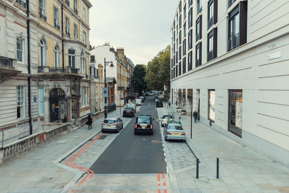 A city street scene showing a narrow, one-way road lined with parked cars on both sides. The cars are a mix of different models and colors, including silver, white, and black, parked parallel along the pavement. On the right, there is a modern, white building with large windows, some with visible reflections, and black metal railings along the sidewalk. On the left, an older, ornate building features detailed stonework, arched windows, decorative balconies, and a black iron gate at the entrance. A few pedestrians are visible walking along the sidewalks, and a person carrying an umbrella is present, suggesting light weather conditions. Trees with green foliage are seen at the end of the street, partly obscured by the buildings, and the overcast sky provides diffuse, natural lighting. The scene is typical of urban environments where private waste collections or rubbish removal services might operate, with the setting reflecting a neighbourhood that could utilize independent rubbish handling options like those offered by Waste Removal Mayfair.