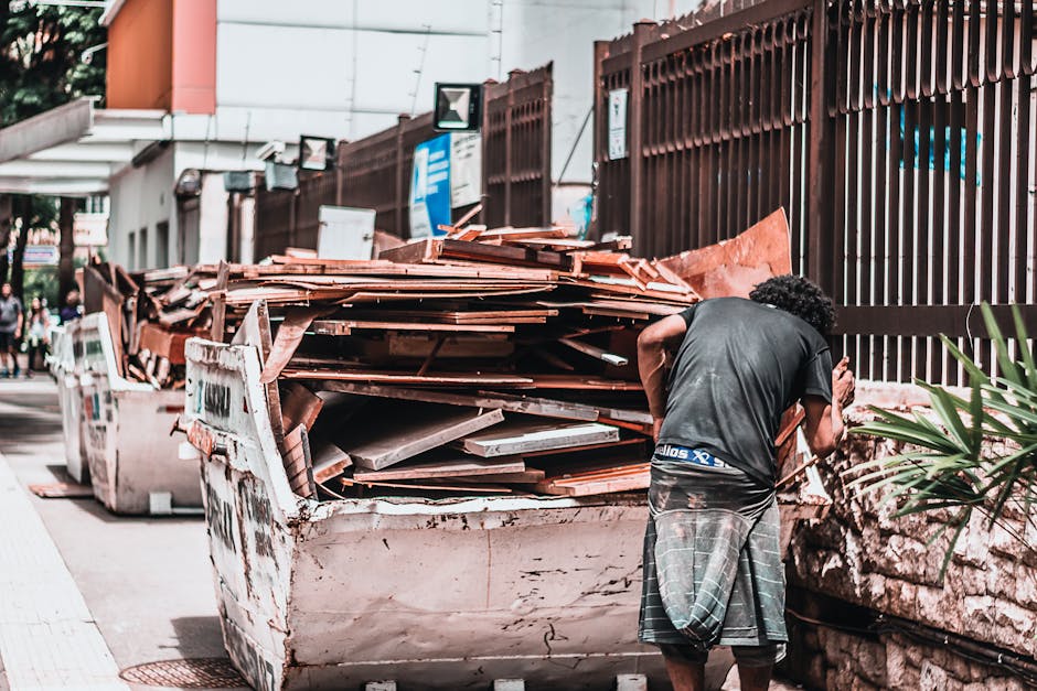 A man with curly dark hair, wearing a black t-shirt and plaid shorts, is standing outdoors on a sidewalk, leaning forward to look into or work around a large, white, steel skip filled with a stack of brown roofing tiles or terracotta roof slates, some of which appear weathered and slightly uneven. The skip is placed next to a high, brown metal fence with vertical bars that enclose a property. In the background, there are other skip containers, some with visible graffiti or dirt, and a few signage and security camera system components mounted on nearby buildings. The scene suggests an ongoing or completed load of building debris, typical of private waste management or alternative rubbish disposal methods, with natural daylight illuminating the scene and casting neutral shadows, indicating clear weather. The environment appears to be an urban or suburban street, with additional pedestrians and buildings faintly visible further in the background, illustrating the context of rubbish removal services such as those offered by Waste Removal Mayfair.