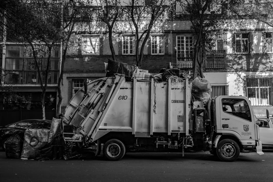 A waste collection truck parked on a city street, featuring an enclosed cab painted in light colour with a distinctive insignia on the door, and a large, rectangular metal body for rubbish storage. The truck is actively engaged in a rubbish collection, with its rear loading mechanism positioned near black trash bags, and various discarded items visible atop the trash container. The environment includes residential buildings with multiple windows and metal security bars, and leafless trees casting shadows on the façade. The scene is captured in black and white, highlighting the textures of the metal surfaces, the crumpled plastic bags, and the urban surroundings, illustrating an organized waste removal operation typical of private waste collection services such as Waste Removal Mayfair. The setting appears to be in a quiet urban neighbourhood, emphasizing the role of independent rubbish collection in alternative waste handling processes for busy city areas.