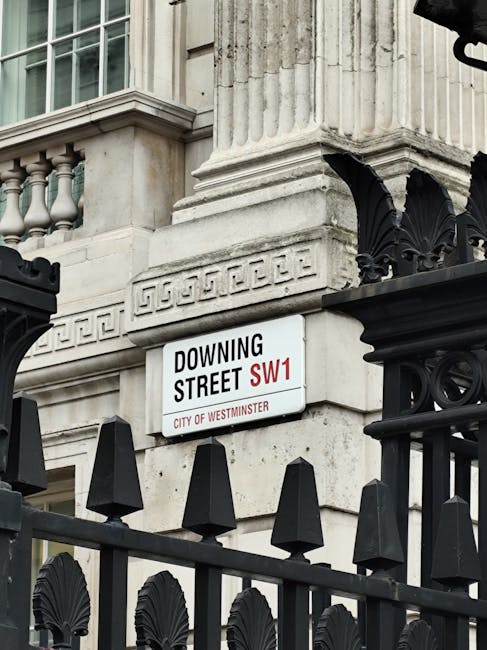 A close-up view of a street sign mounted on a building corner in Mayfair, London, displaying the street name 'Downing Street' with the postal code 'SW1' and the label 'City of Westminster' beneath. The building features classical architectural details, including a large fluted column with decorative molding, made of light-colored stone or concrete. In the foreground, a black wrought iron gate or fence with pointed finials and ornate patterns partially obstructs the view of the sign. The architectural elements convey an elegant, historic style typical of central London buildings. The lighting appears natural, with soft shadows indicating an outdoor setting during daytime. The setting suggests an area where private or official properties are situated, and the presence of the fence hints at aspects of security or private access, which may relate to the context of private waste handling or restricted entry often associated with such premises. Waste Removal Mayfair would handle rubbish collection for similar properties, ensuring proper and compliant disposal of household waste in this prestigious district.