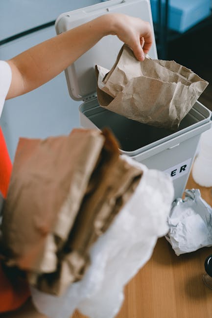 A person inserts a crumpled brown paper bag into a white bin designated for waste removal, positioned on a wooden surface. The bin has a partially open lid and is clearly marked for rubbish collection. In the foreground, several other paper bags are held, with their edges visible, indicating they are ready for disposal. The scene captures an indoor environment, suggesting a household or office setting, with natural or artificial light illuminating the scene evenly. The bags appear to be made of kraft paper with a textured surface, typical for disposable rubbish. The focus is on the process of waste sorting, aligning with private waste handling practices that may be managed by services like Waste Removal Mayfair, emphasizing proper disposal of household or commercial waste through independent collection methods. The background includes some additional clutter, possibly other waste items or packing materials, reinforcing the theme of waste management and rubbish removal services.